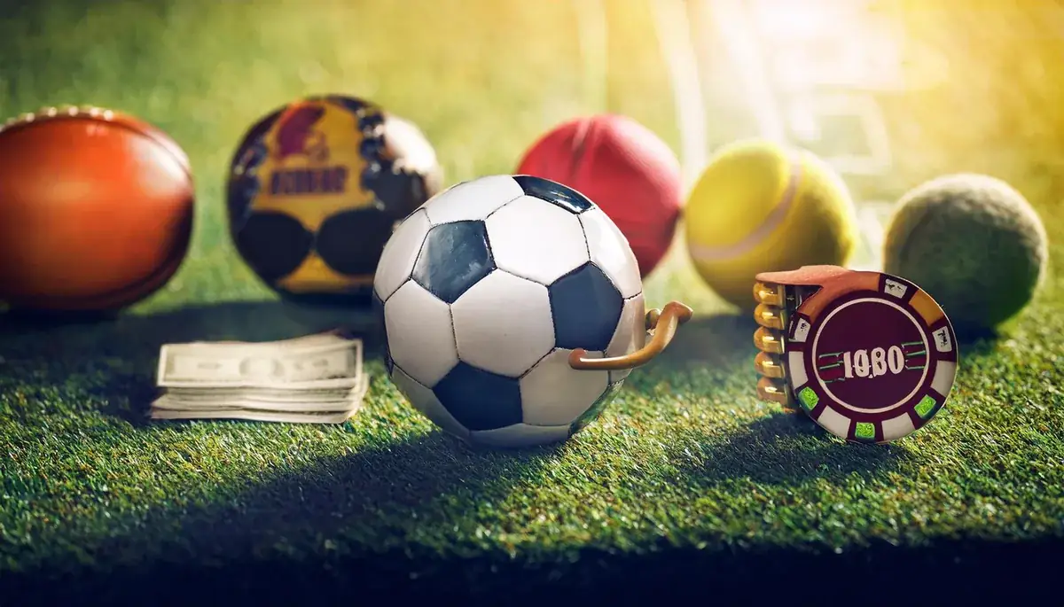 A football in sharp focus on green grass, with a rugby ball, a cricket ball, tennis balls, a folded stack of banknotes and a red casino chip arranged around it